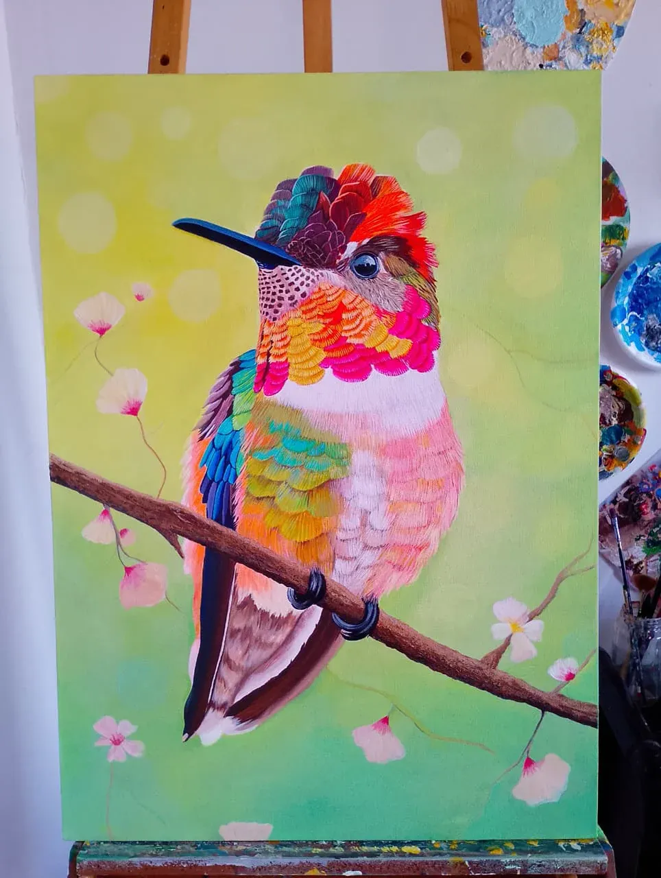 Colibrí de plumas multicolor posado en una rama con flores rosadas sobre fondo verde